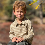 A young boy with curly light brown hair sits outdoors on a weathered wooden fence. He is wearing a beige and tan horizontally striped long-sleeve henley shirt and dark gray jeans.