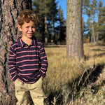 A young boy with curly light brown hair stands outdoors leaning against a tree trunk in a grassy field. He is wearing a long-sleeve polo shirt with navy, burgundy, and white horizontal stripes, paired with khaki pants.