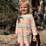 A young girl with blonde hair stands outdoors in a wooded area, wearing a long-sleeve striped tunic dress with pastel peach, blue, and green stripes on an off-white base, paired with pink leggings.
