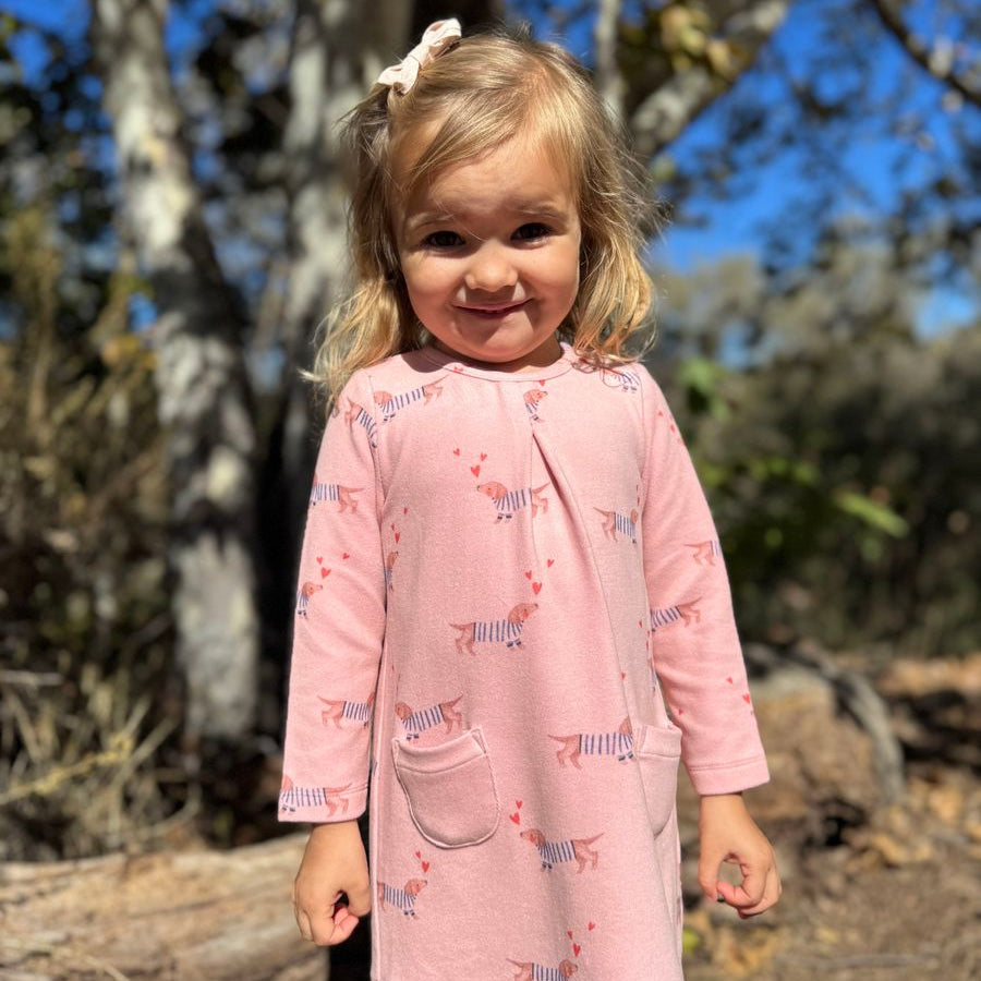 A young toddler girl with blonde hair wearing a pink long-sleeve tunic dress with a zebra print, standing outdoors in front of trees and blue sky.