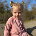 A toddler girl with blonde pigtails sits outdoors wearing a pink long-sleeve romper with a small animal print. The background shows a blurred natural setting with blue sky and trees.