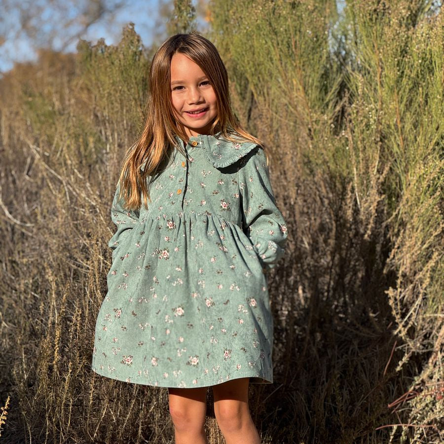 Young girl in a light green floral dress standing in a natural setting with bushes and blue sky.