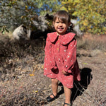 A young girl smiling outdoors in a rust-red long-sleeve dress with a white floral pattern, standing on dry grass with trees in the background.