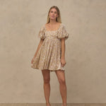 A woman modeling a short puff-sleeve dress with a beige and white floral pattern, standing against a textured beige studio backdrop.
