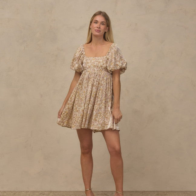 A woman modeling a short puff-sleeve dress with a beige and white floral pattern, standing against a textured beige studio backdrop.