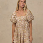 A woman modeling a short puff-sleeve dress with a beige and brown floral pattern against a textured beige wall.