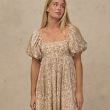A woman modeling a short puff-sleeve dress with a beige and brown floral pattern against a textured beige wall.