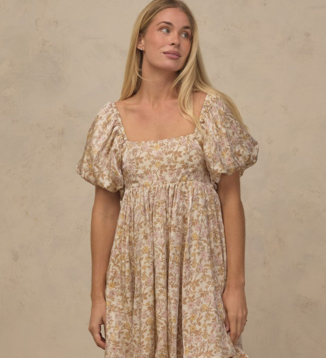 A woman modeling a short puff-sleeve dress with a beige and brown floral pattern against a textured beige wall.