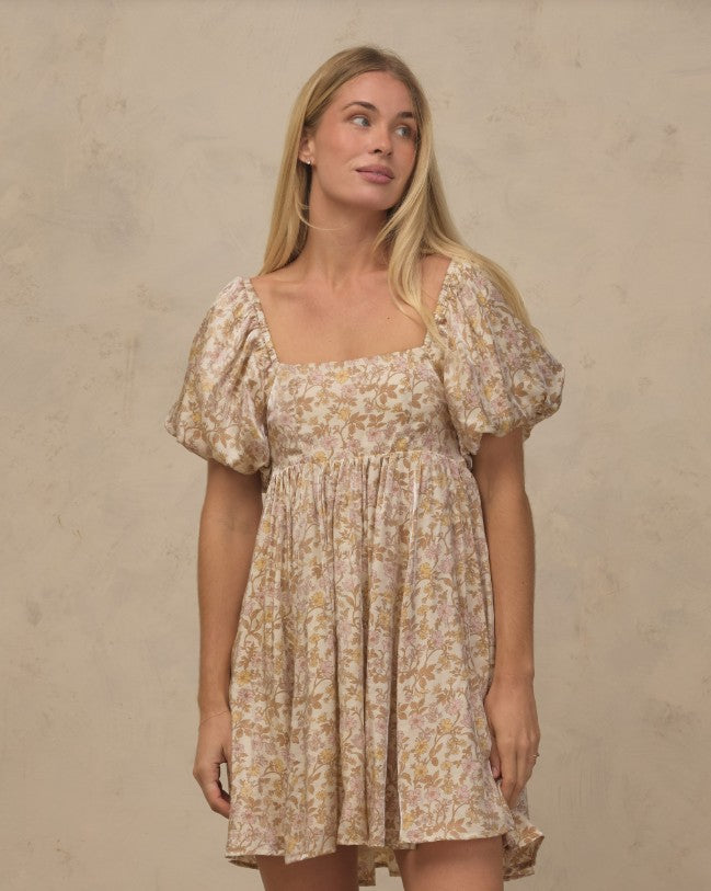 A woman modeling a short puff-sleeve dress with a beige and brown floral pattern against a textured beige wall.