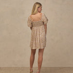 A woman stands with her back to the camera wearing a short puff-sleeve mini dress with a beige and yellow floral pattern and a smocked bodice, paired with beige high-heeled sandals, against a textured beige studio backdrop.