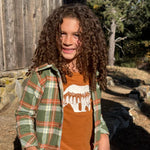A young girl with long, curly brown hair stands outdoors in front of a rustic wooden barn wall. She is wearing a green and orange plaid flannel shirt over an orange crewneck t-shirt with a white bear graphic, paired with blue jeans.