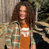 A young girl with long, curly brown hair stands outdoors in front of a rustic wooden barn wall. She is wearing a green and orange plaid flannel shirt over an orange crewneck t-shirt with a white bear graphic, paired with blue jeans.