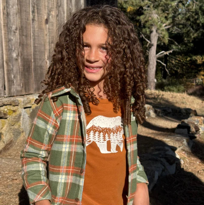 A young girl with long, curly brown hair stands outdoors in front of a rustic wooden barn wall. She is wearing a green and orange plaid flannel shirt over an orange crewneck t-shirt with a white bear graphic, paired with blue jeans.