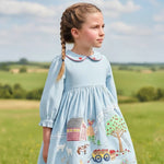 A young girl with braided hair wearing a light blue long-sleeve dress with embroidered farm-themed motifs on the skirt, standing outdoors in a grassy field under a cloudy sky.