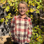 A young boy smiling outdoors in front of autumn trees, wearing a long-sleeve buffalo check plaid button-down shirt in red, beige, and brown tones.