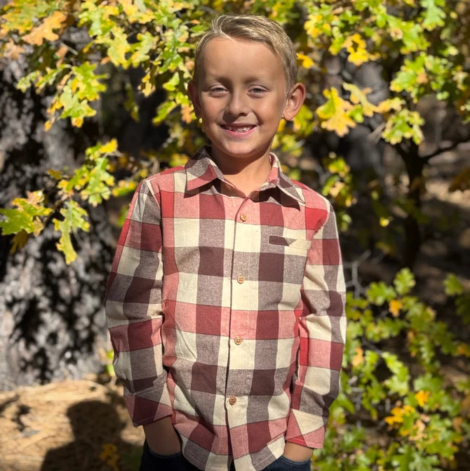 A young boy smiling outdoors in front of autumn trees, wearing a long-sleeve buffalo check plaid button-down shirt in red, beige, and brown tones.