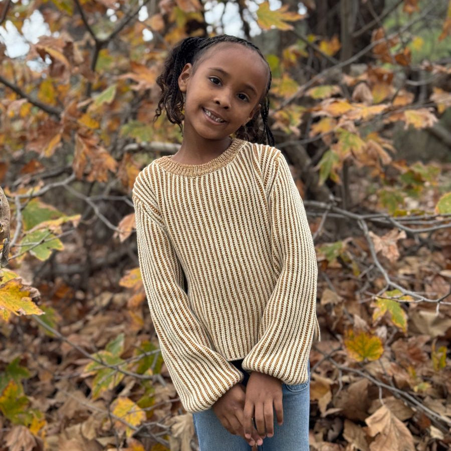 A young girl with braided hair stands outdoors in a wooded area with fallen leaves, wearing a beige and cream vertically striped knit sweater and blue jeans.