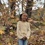 A young girl with dark curly hair stands outdoors in front of autumn trees, wearing a cream ribbed knit sweater and blue jeans.