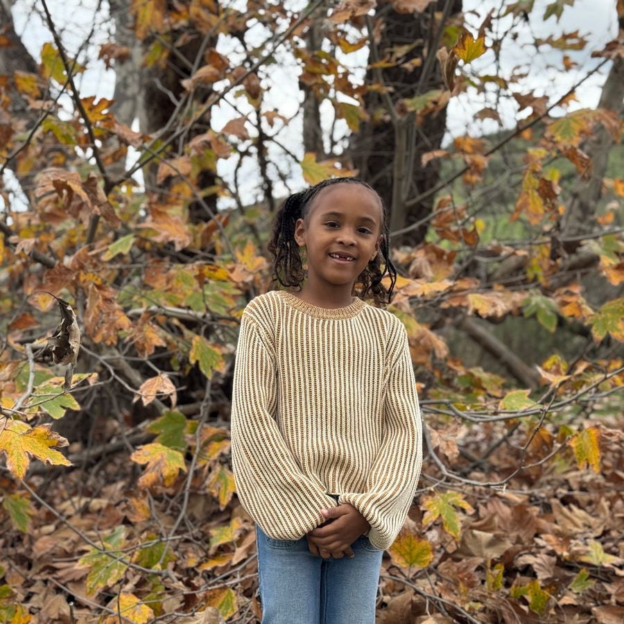 A young girl with dark curly hair stands outdoors in front of autumn trees, wearing a cream ribbed knit sweater and blue jeans.