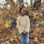 A young girl with braided hair stands outdoors in a wooded area covered with fallen leaves, wearing a cream and tan vertically striped long-sleeve knit sweater and blue jeans.