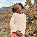 Young girl in a beige sweater and pink skirt standing in an autumn landscape with trees and mountains.