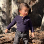 A toddler standing outdoors in a wooded area, wearing a navy long-sleeve henley onesie with brown buttons and dark gray jeans.