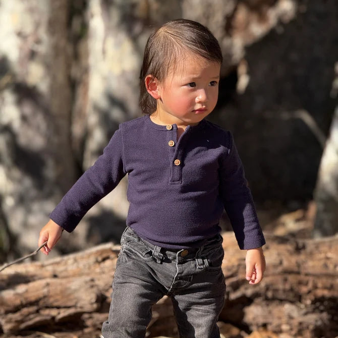 A toddler standing outdoors in a wooded area, wearing a navy long-sleeve henley onesie with brown buttons and dark gray jeans.