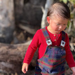 A toddler wearing a red long-sleeve henley shirt and navy blue plaid overalls, standing outdoors in a wooded setting.