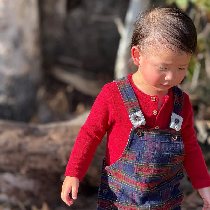 A toddler wearing a red long-sleeve henley shirt and navy blue plaid overalls, standing outdoors in a wooded setting.