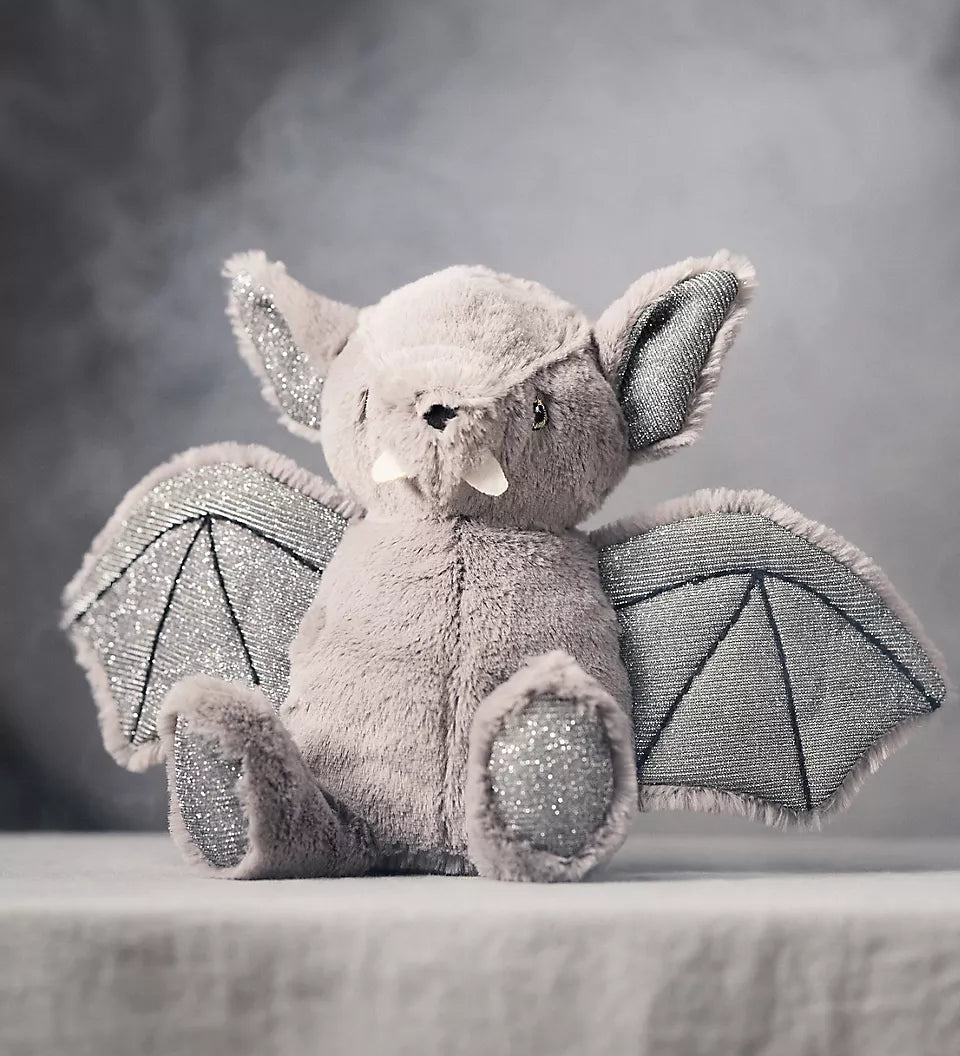 A plush bat toy sitting on a draped fabric surface against a smoky gray studio background. The toy is light gray with darker gray wings and small white accents on the ears and snout.