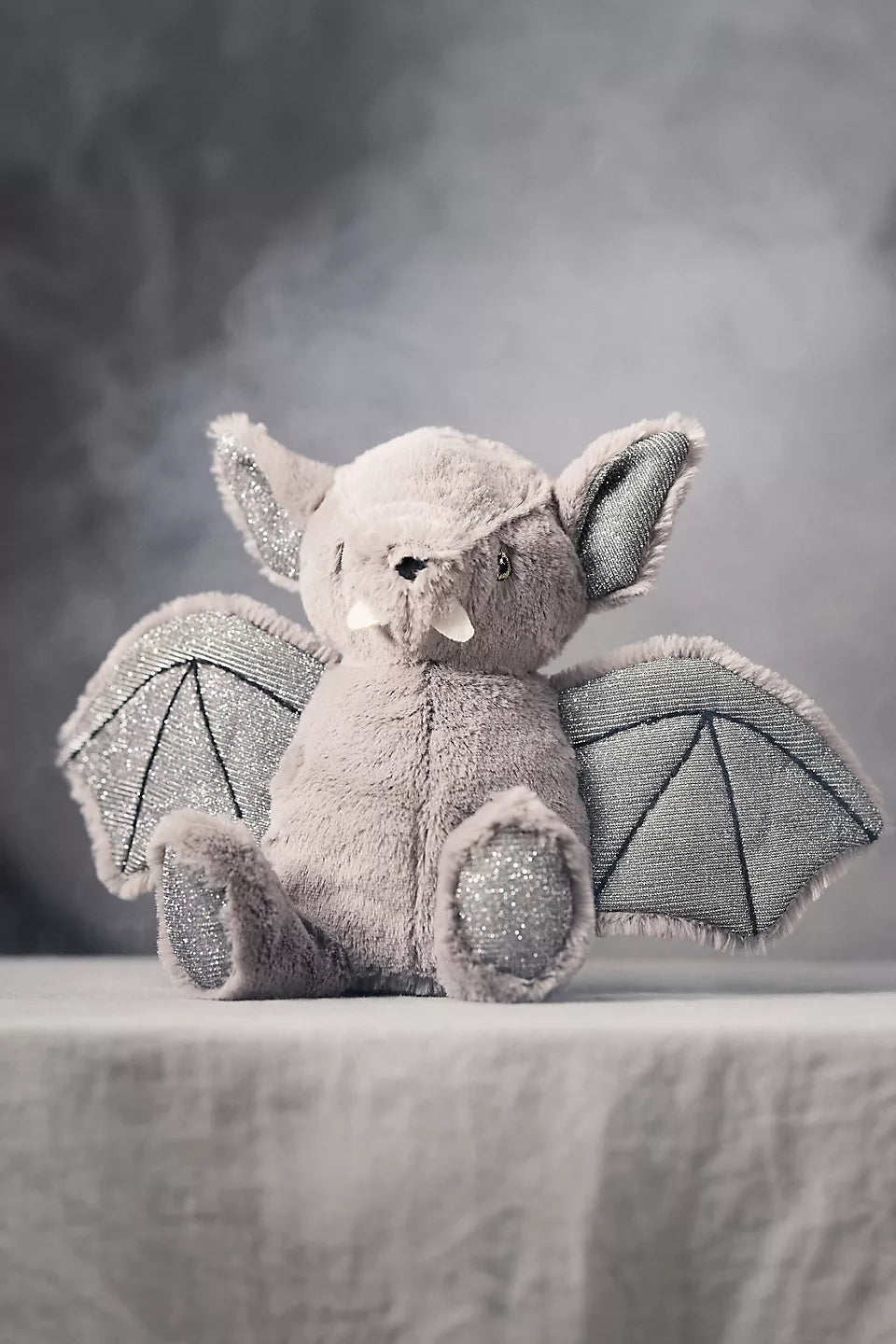 A plush bat toy sitting on a draped fabric surface against a smoky gray studio background. The toy is light gray with darker gray wings and small white accents on the ears and snout.