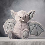 A plush bat toy sitting on a draped fabric surface against a smoky gray studio background. The toy is light gray with darker gray wings and small white accents on the ears and snout.