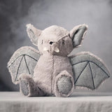 A plush bat toy sitting on a draped fabric surface against a smoky gray studio background. The toy is light gray with darker gray wings and small white accents on the ears and snout.