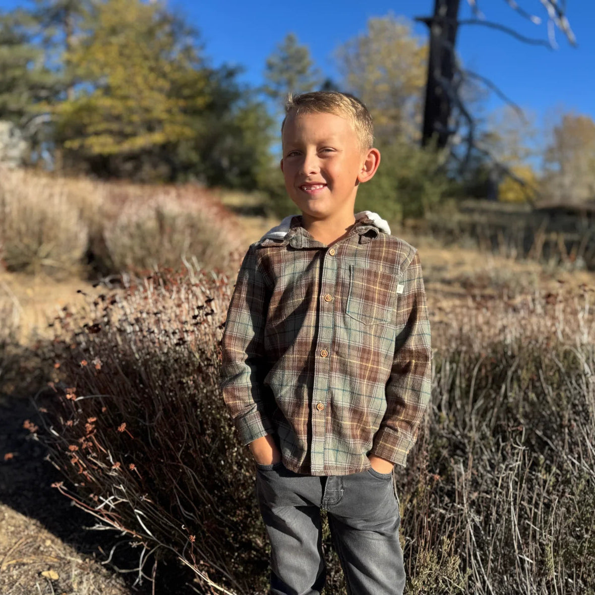 A young boy smiling outdoors in a brown and beige plaid hooded button-up shirt and gray pants, standing in a sunny, brush-filled landscape.