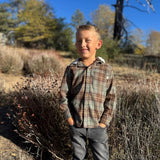 A young boy smiling outdoors in a brown and beige plaid hooded button-up shirt and gray pants, standing in a sunny, brush-filled landscape.