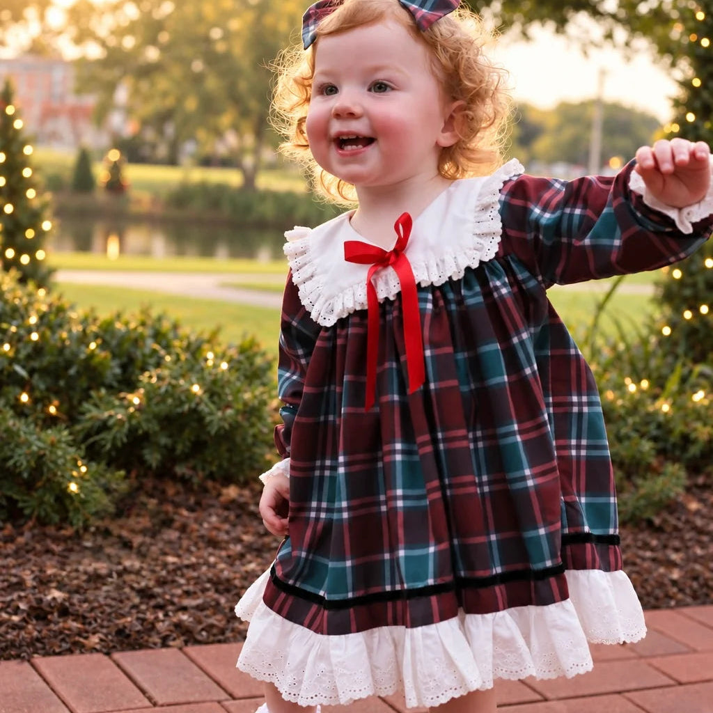 A toddler girl wearing a festive holiday plaid dress with white ruffles and a red bow, standing on a brick path in a garden at sunset.