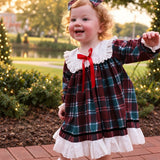 A toddler girl wearing a festive holiday plaid dress with white ruffles and a red bow, standing on a brick path in a garden at sunset.