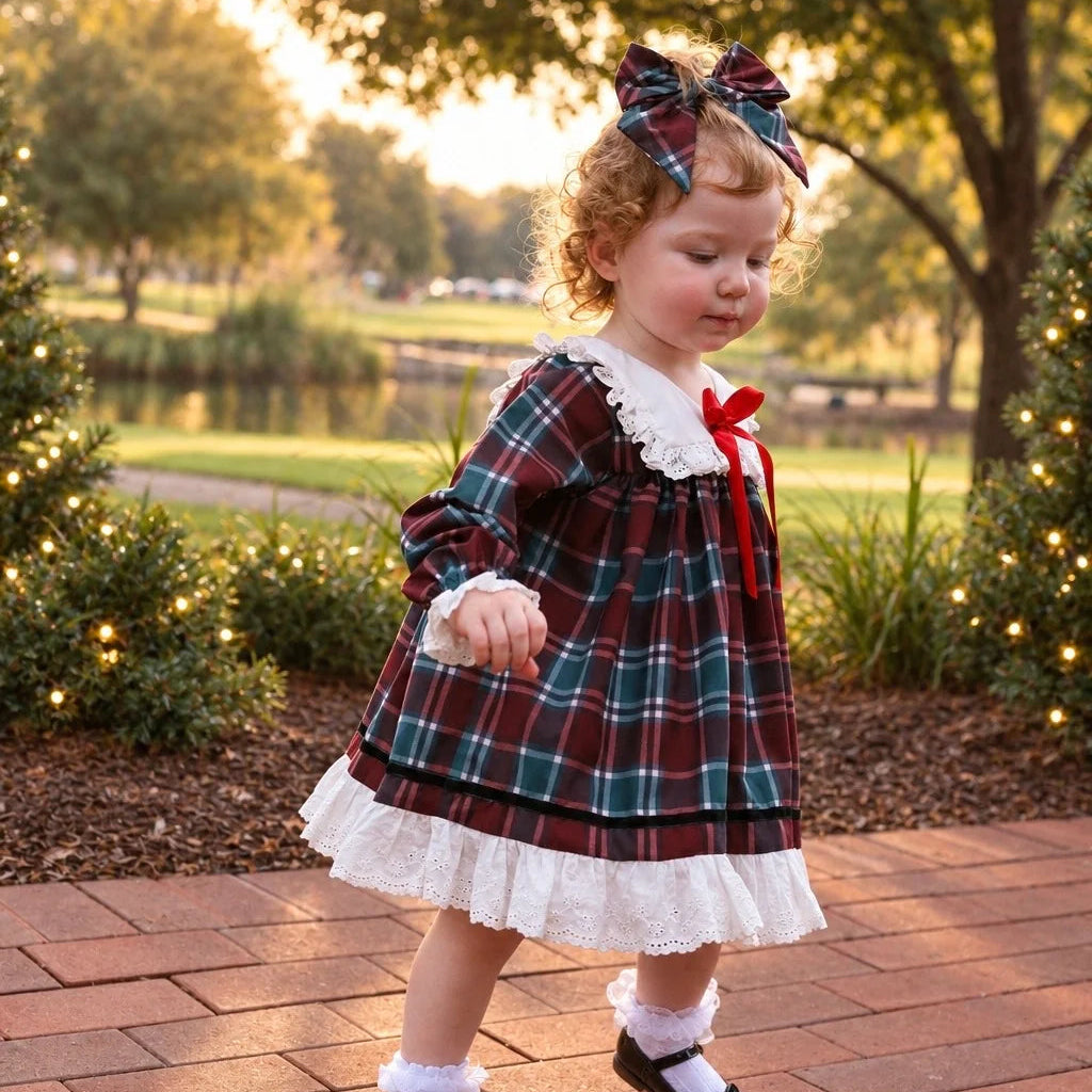 A toddler girl wearing a red and green plaid dress with white lace trim, a red bow at the collar, and a matching plaid bow headband, standing on a brick path in an outdoor park setting at sunset.