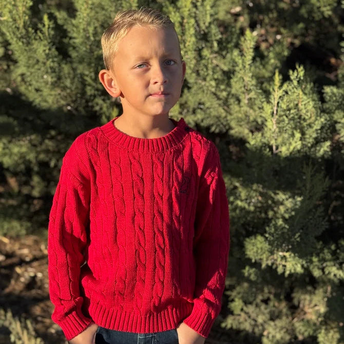 A young boy wearing a bright red cable-knit sweater and dark jeans, standing outdoors in front of green evergreen trees.