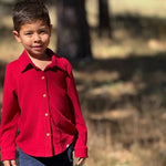 A young boy wearing a bright red long-sleeve button-up shirt and navy blue pants, standing outdoors in a sunny, dry grassy field.