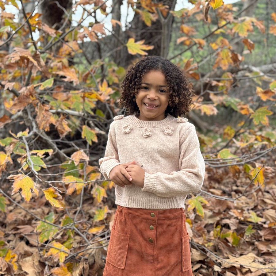 A young girl with curly dark hair stands outdoors in a wooded area with autumn leaves, wearing a cream knit sweater with decorative pom-pom details on the chest and a rust-orange skirt.