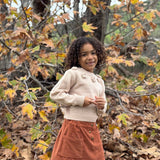 A young girl with curly dark hair stands outdoors in an autumn forest setting, wearing a cream ribbed knit sweater with decorative buttons and a rust-orange corduroy skirt.