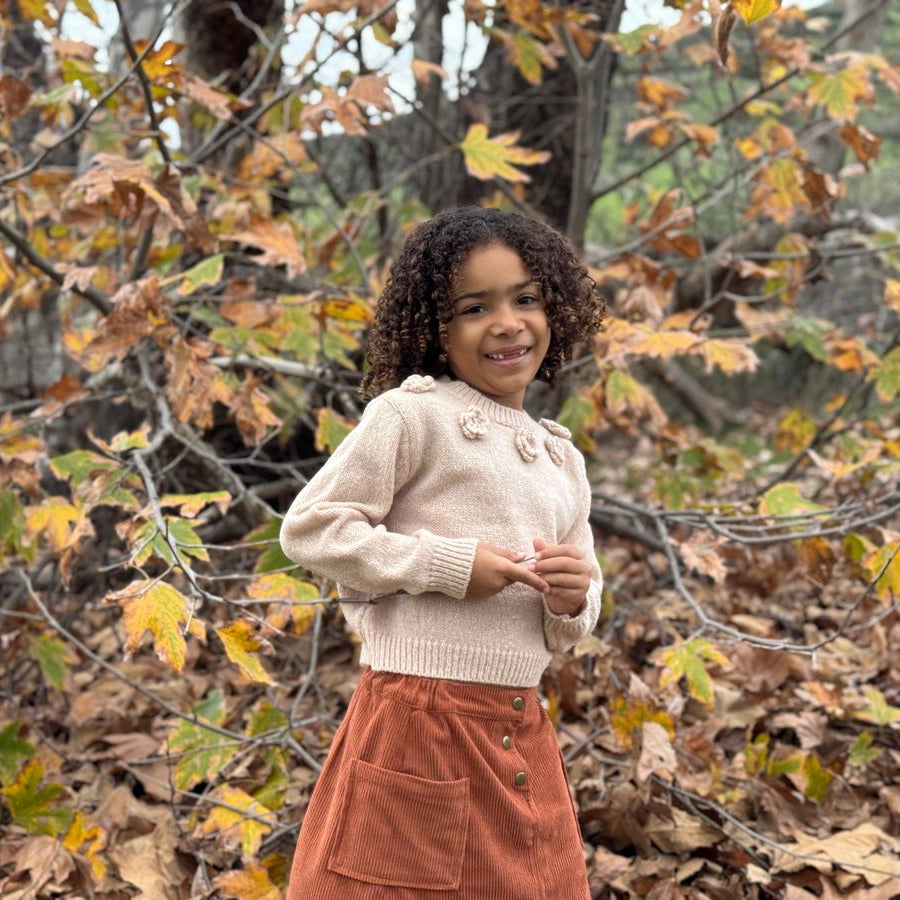 A young girl with curly dark hair stands outdoors in an autumn forest setting, wearing a cream ribbed knit sweater with decorative buttons and a rust-orange corduroy skirt.