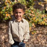 A young boy with curly brown hair stands outdoors in a field of dry leaves and autumn foliage, wearing a beige and brown checkered long-sleeve button-up shirt and dark blue jeans.