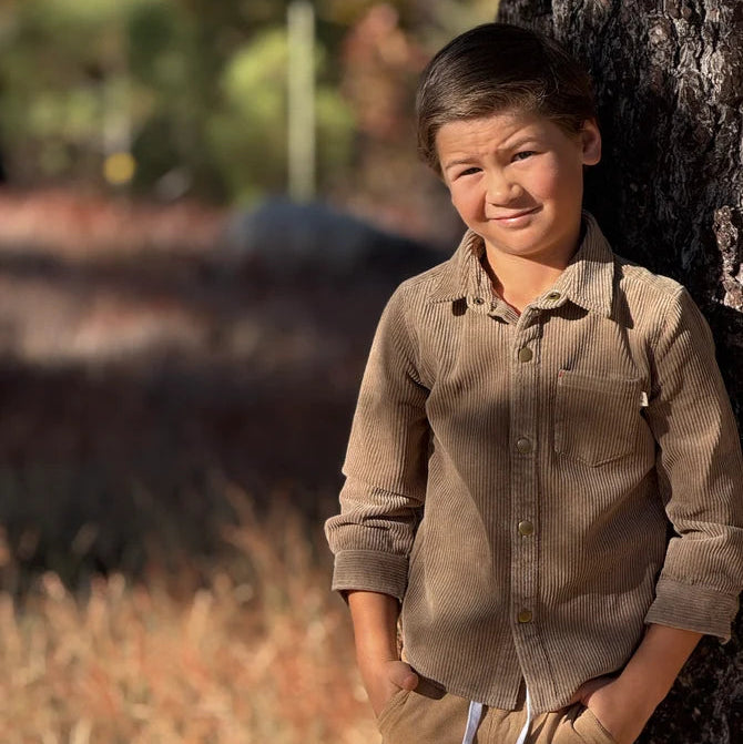 Young boy in a brown shirt and pants standing next to a tree in a natural setting