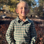 A young boy with blond hair stands outdoors wearing a green and beige plaid long-sleeve button-up shirt and blue jeans, with a blurred natural background of trees and brush.