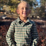 A young boy with blond hair stands outdoors wearing a green and beige plaid long-sleeve button-up shirt and blue jeans, with a blurred natural background of trees and brush.