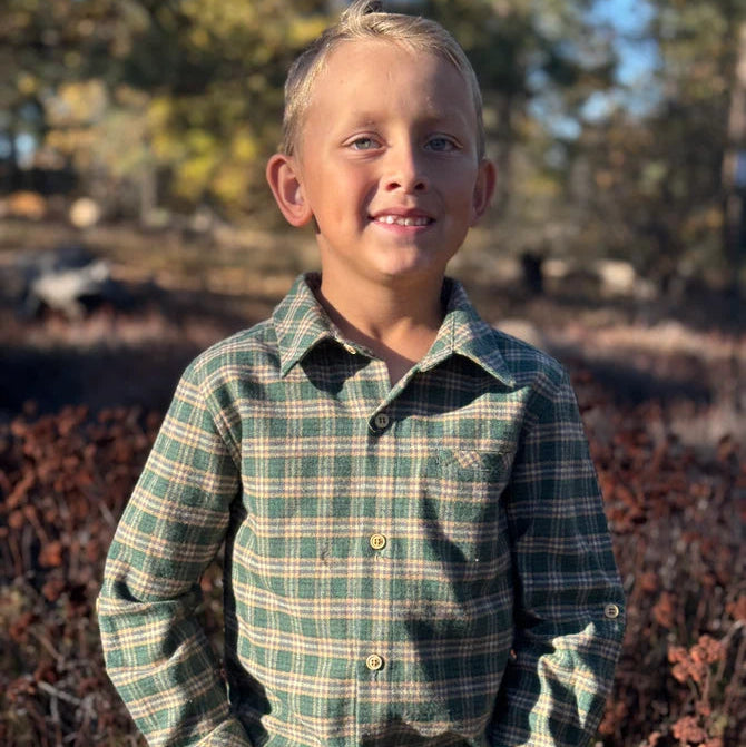 A young boy with blond hair stands outdoors wearing a green and beige plaid long-sleeve button-up shirt and blue jeans, with a blurred natural background of trees and brush.