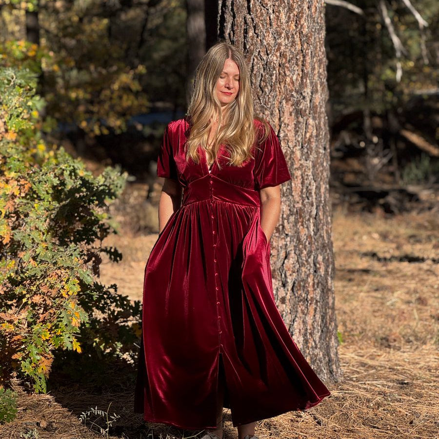 A woman wearing a long burgundy velvet midi dress with short sleeves and a buttoned waist stands outdoors next to a large tree trunk in a wooded setting.