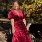 A woman wearing a deep burgundy velvet midi dress with short puff sleeves and a V-neckline, standing outdoors in front of a blurred background of autumn trees.